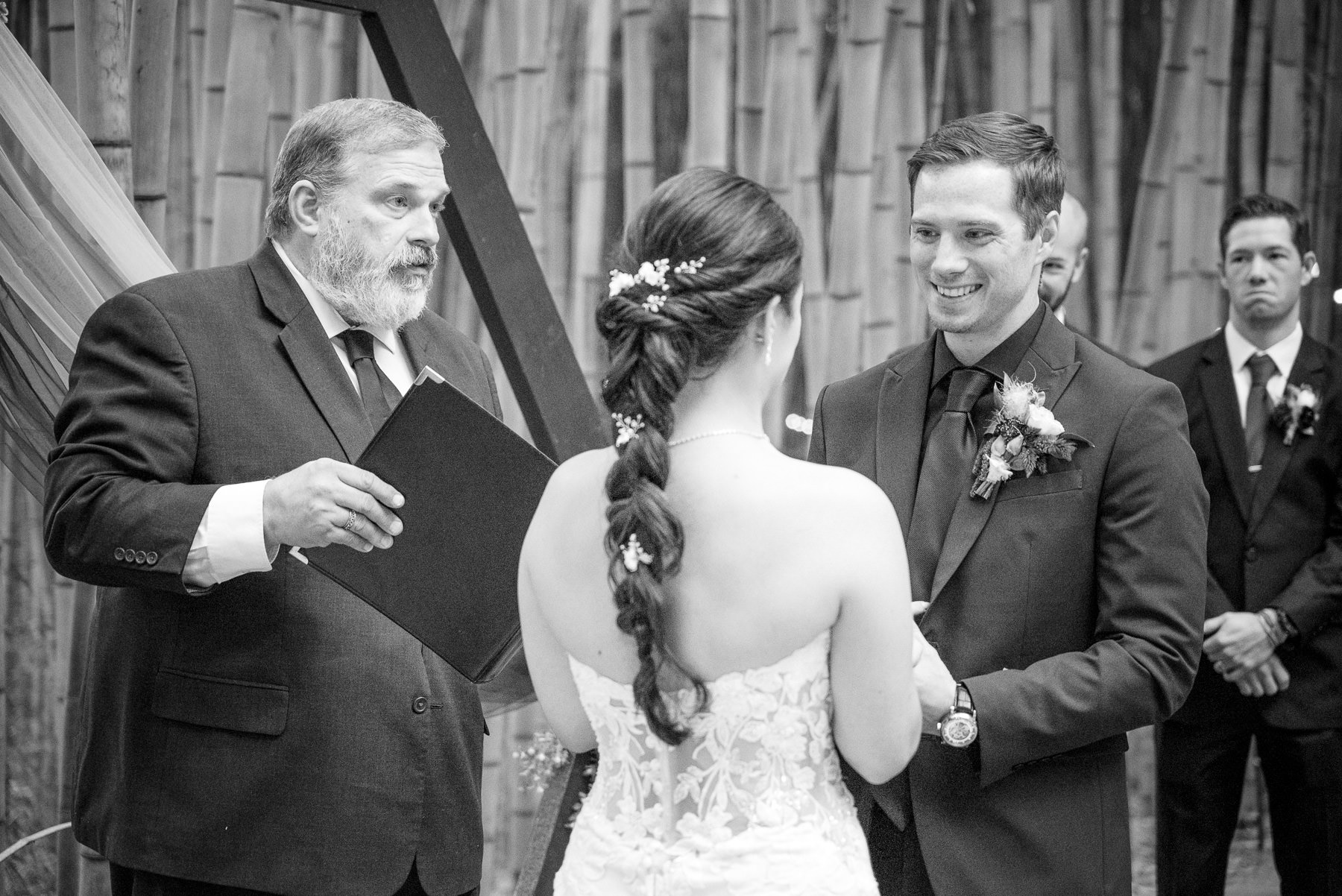 Bride and groom exchange vows during their outdoor wedding ceremony at Camelot Meadows in Charlotte, NC, smiling as they hold hands beneath a modern floral arch surrounded by a stunning bamboo grove. Captured by Charlotte wedding photographers DeLong Photography.