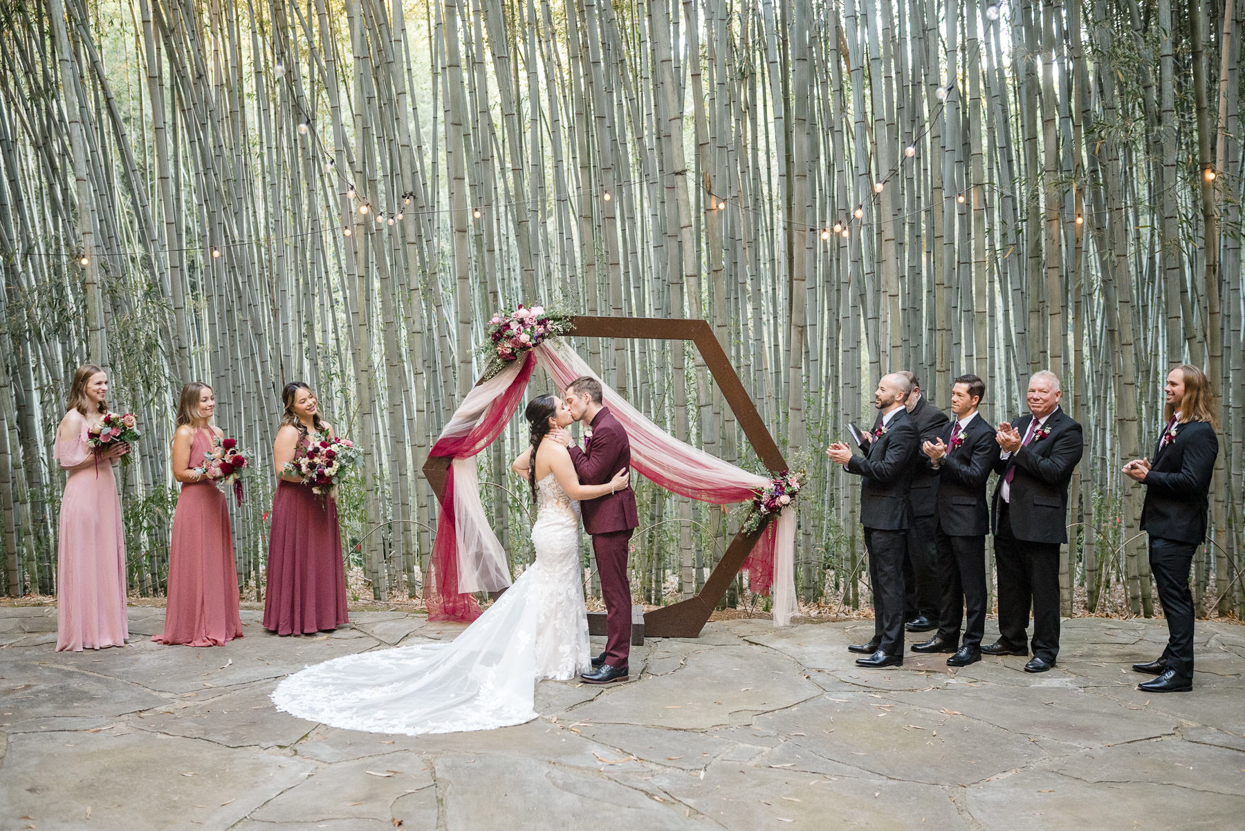 Bride and groom share their first kiss as newlyweds during an outdoor wedding ceremony at Camelot Meadows in Charlotte, NC, beneath a modern hexagon arch with burgundy and blush florals, surrounded by their wedding party in a breathtaking bamboo grove. Captured by Charlotte wedding photographers DeLong Photography.
