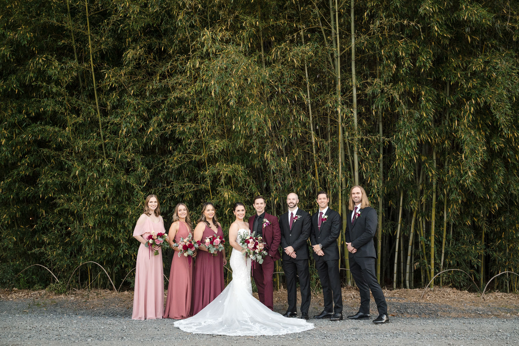 Bridal party portrait at Camelot Meadows wedding venue in Charlotte, NC, with the bride and groom standing alongside bridesmaids and groomsmen in front of a towering bamboo forest. Captured by DeLong Photography, this elegant outdoor wedding showcases romantic burgundy florals, modern bridal party attire, and timeless wedding photography for Charlotte couples.