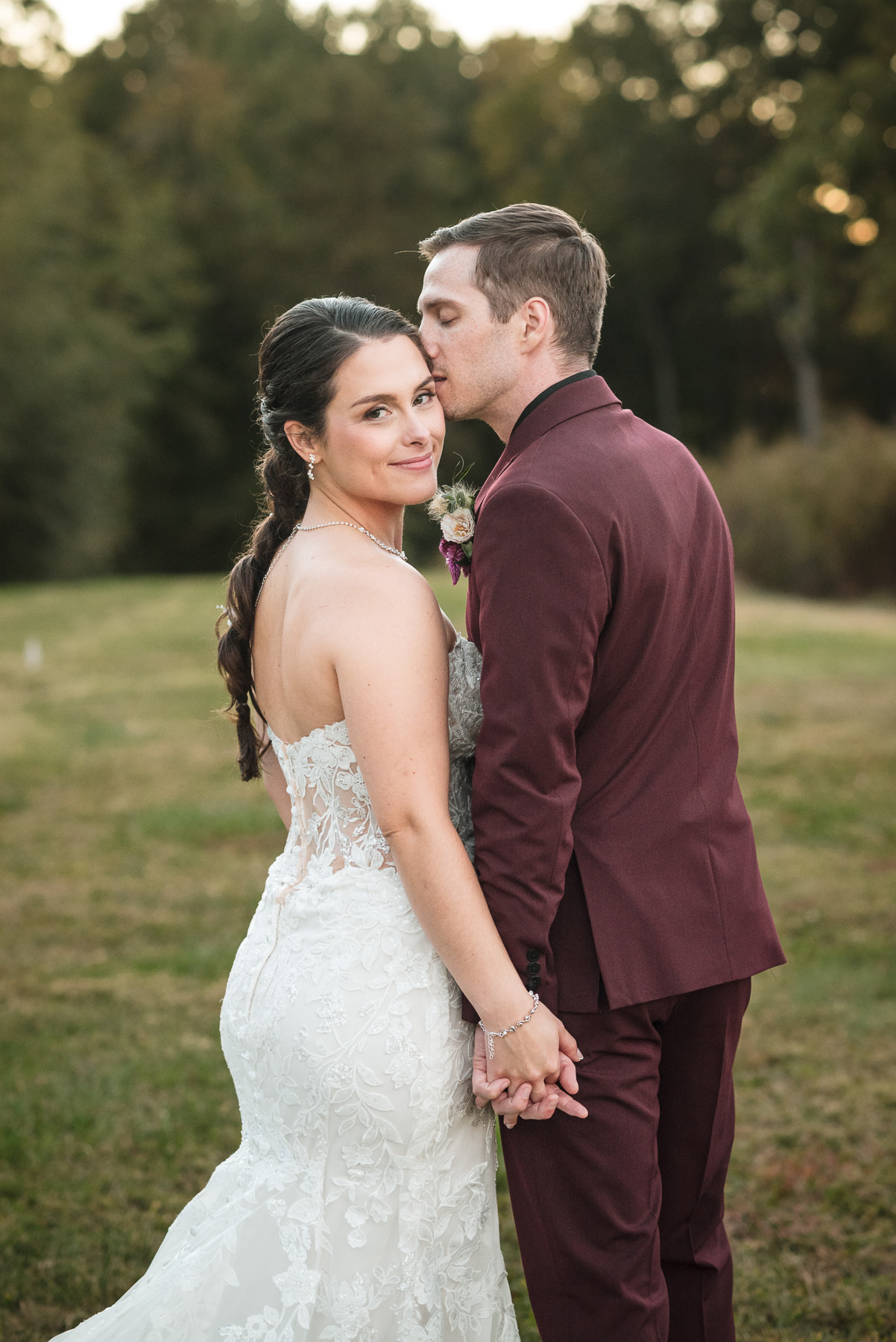 Bride and groom hold hands during romantic sunset portraits at Camelot Meadows in Belmont, North Carolina, as the groom kisses the bride's forehead. Captured by DeLong Photography, this timeless outdoor wedding portrait highlights the venue's natural beauty and elegant Charlotte wedding photography.