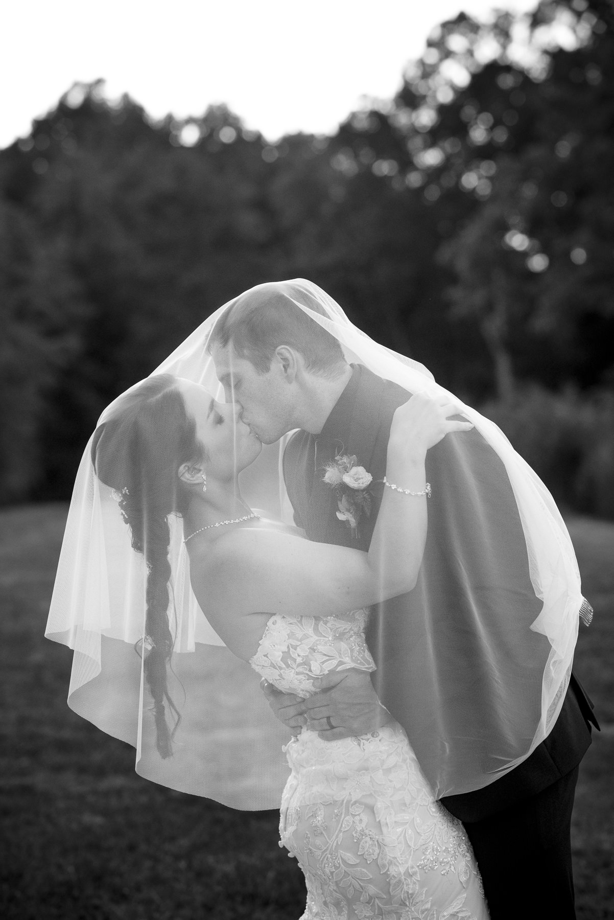 Black-and-white portrait of a bride and groom sharing a kiss beneath the bride's veil during sunset portraits at Camelot Meadows in Belmont, North Carolina. Captured by DeLong Photography, this romantic outdoor wedding image showcases timeless Charlotte wedding photography at one of the area's most scenic wedding venues.