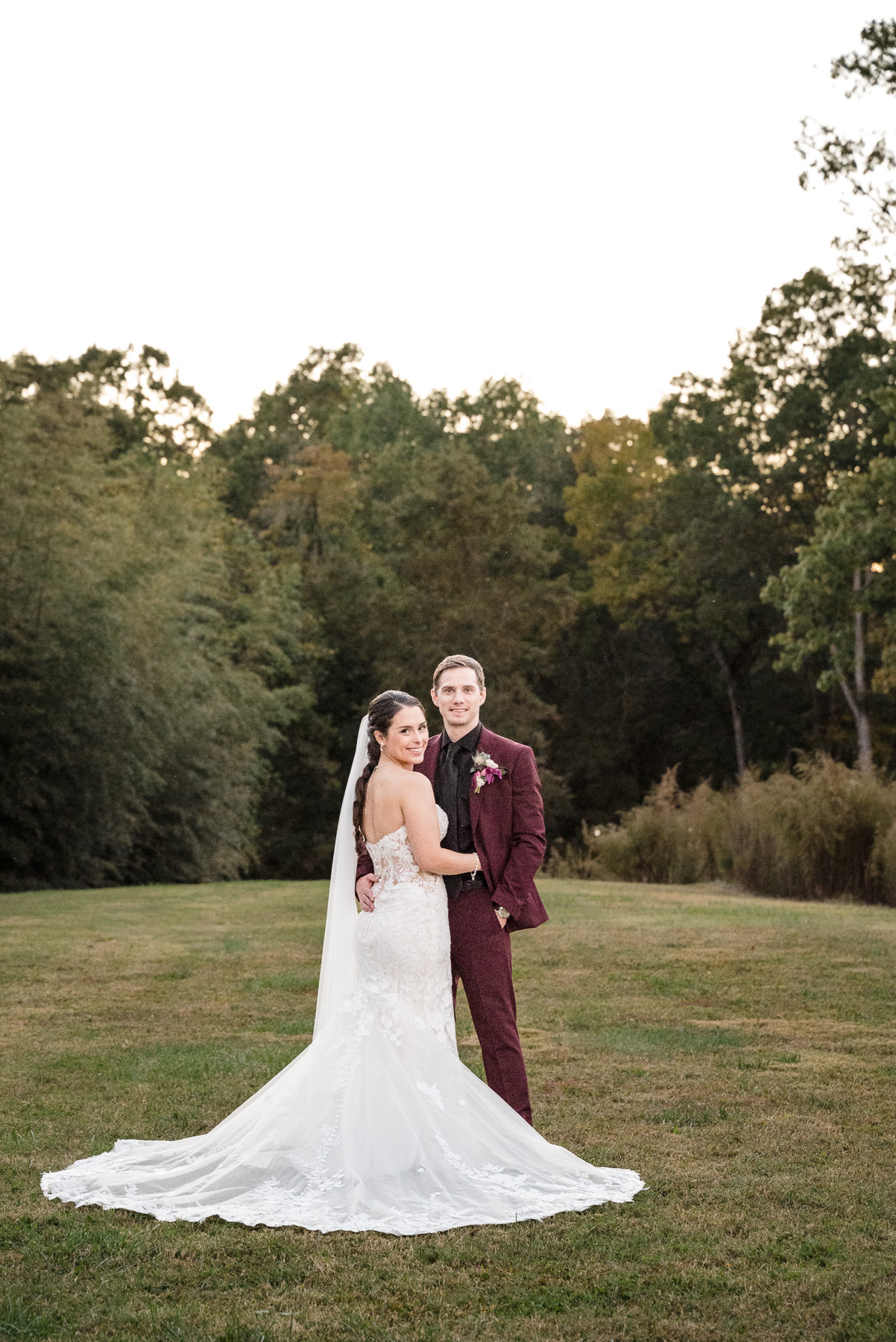 Bride and groom embrace during golden hour portraits at Camelot Meadows in Belmont, North Carolina, with the bride's lace train flowing across the lawn. Captured by DeLong Photography, this elegant outdoor wedding portrait highlights timeless Charlotte wedding photography at one of the region's most beautiful wedding venues.