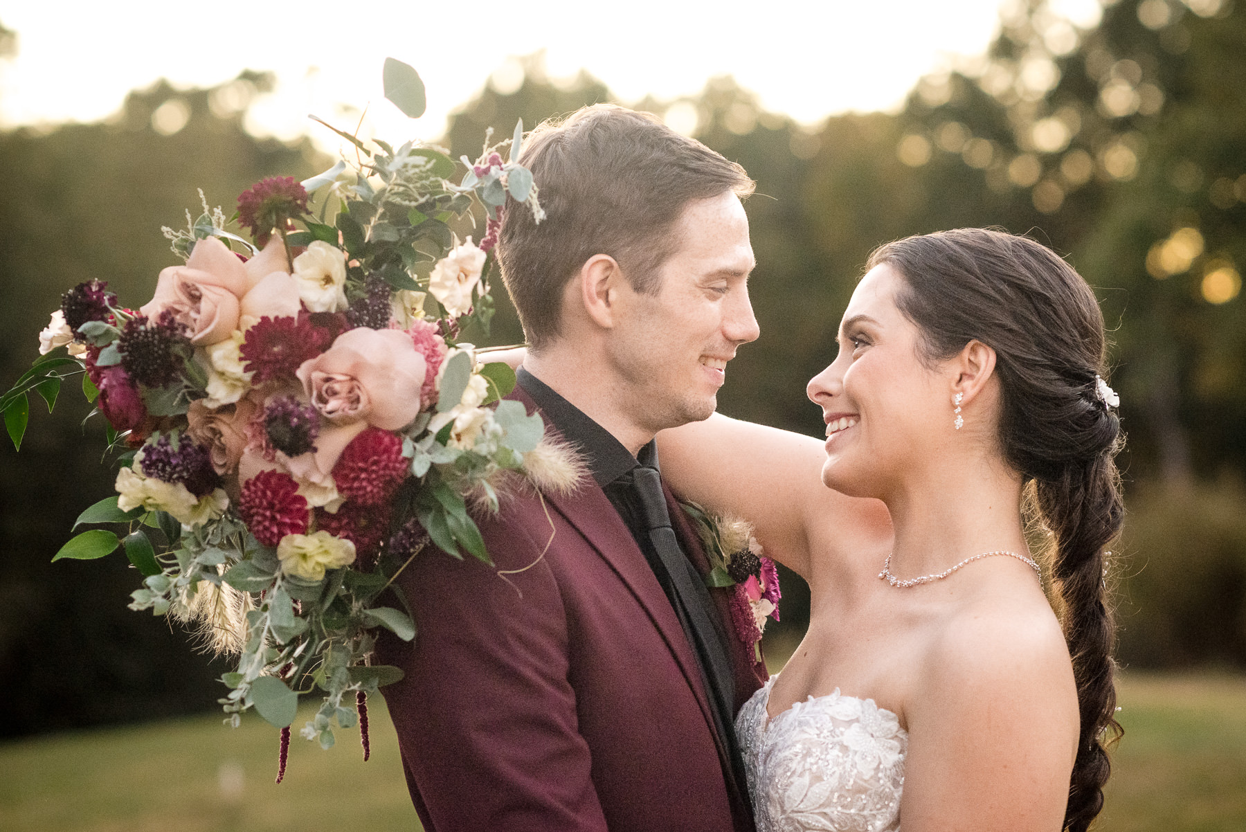 Bride and groom smile at each other during golden hour portraits at Camelot Meadows in Belmont, North Carolina, with the bride's burgundy, blush, and ivory bouquet glowing in the sunset light. Captured by DeLong Photography, this romantic outdoor wedding portrait showcases timeless Charlotte wedding photography and elegant floral details.