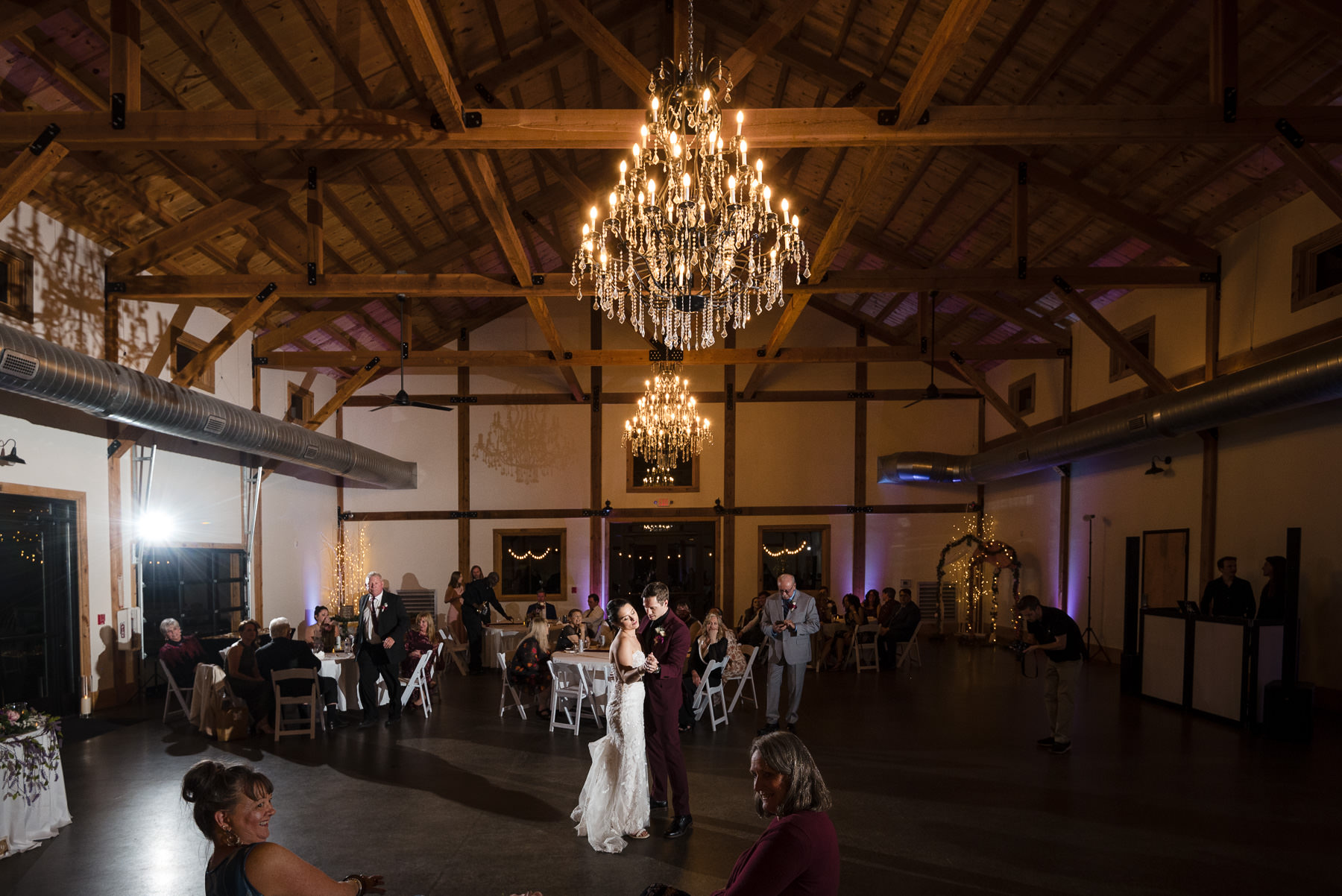 Bride and groom share their first dance beneath sparkling crystal chandeliers inside the reception hall at Camelot Meadows in Belmont, North Carolina. Captured by DeLong Photography, this elegant wedding reception image showcases the venue's stunning timber-frame architecture and timeless Charlotte wedding photography.
