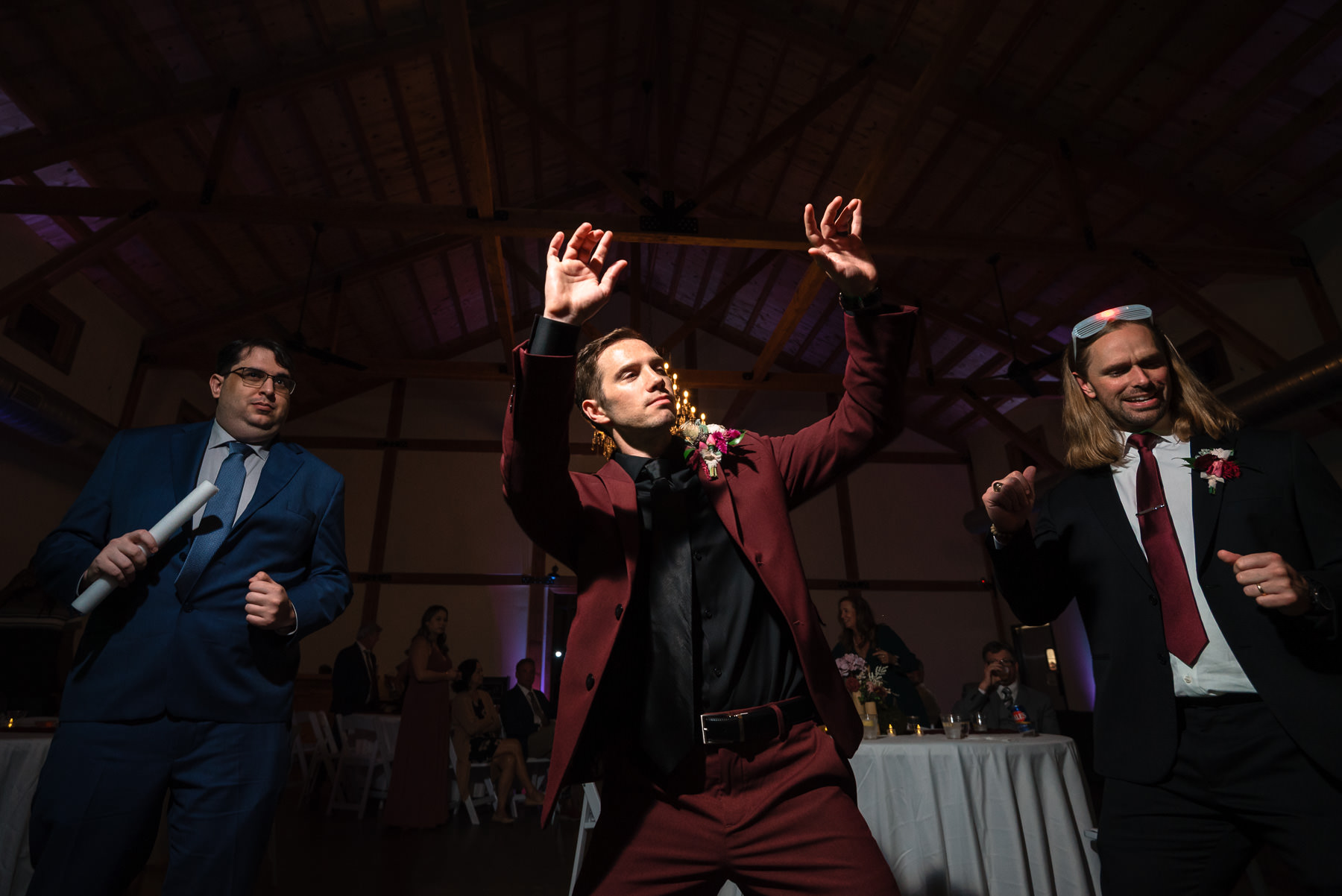 Groom in a burgundy suit dances with friends during the wedding reception at Camelot Meadows in North Carolina. This lively reception photo captures the fun atmosphere inside the elegant timber-frame venue, showcasing the celebration, dance floor, and candid moments photographed by Charlotte wedding photographers DeLong Photography.