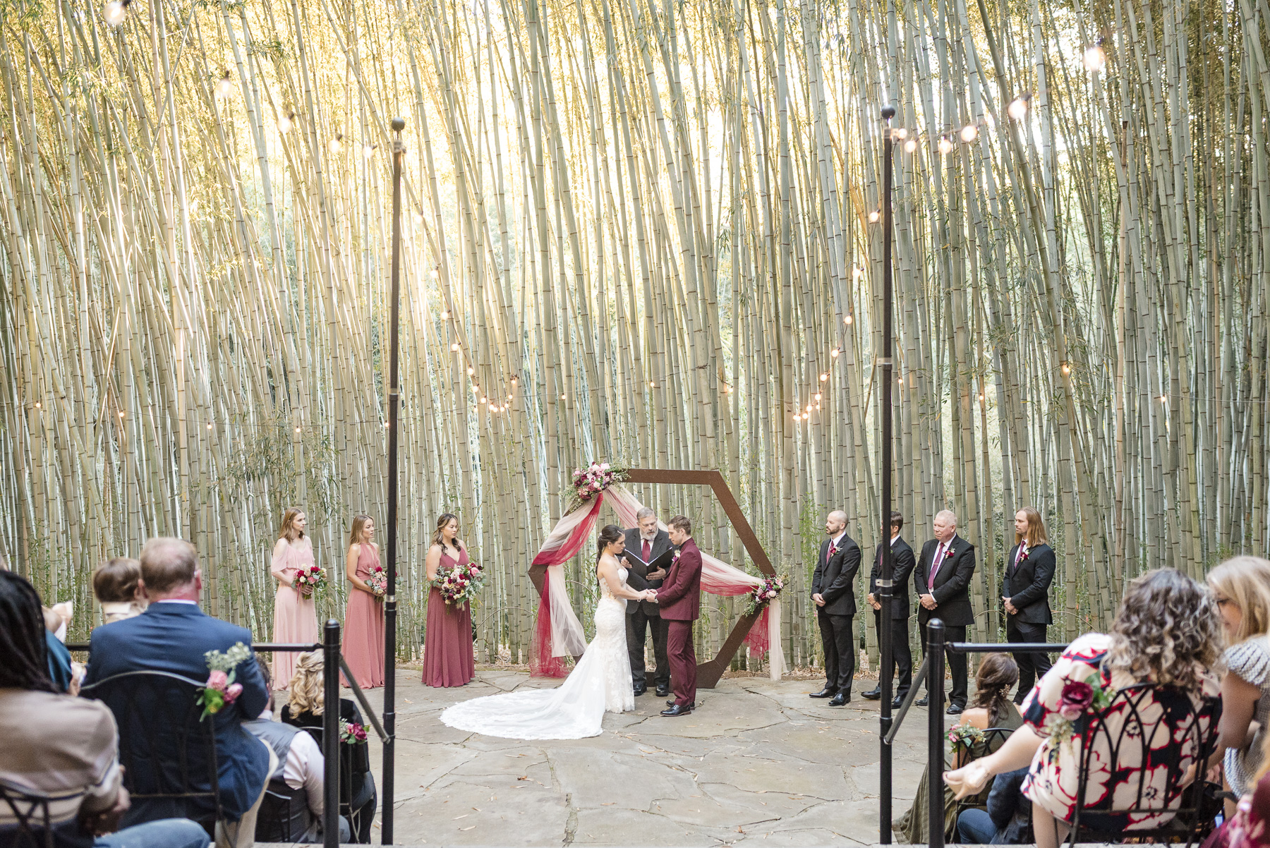 Bride and groom exchange vows during an outdoor wedding ceremony beneath towering bamboo at Camelot Meadows in Charlotte, NC, surrounded by family, bridal party, and romantic string lights. Timeless ceremony photography by Charlotte wedding photographers DeLong Photography.