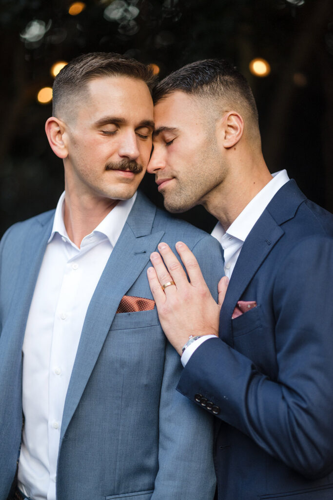 Two grooms share an intimate portrait at McGill Rose Garden in Charlotte, NC, wearing blue suits during their elegant wedding celebration.