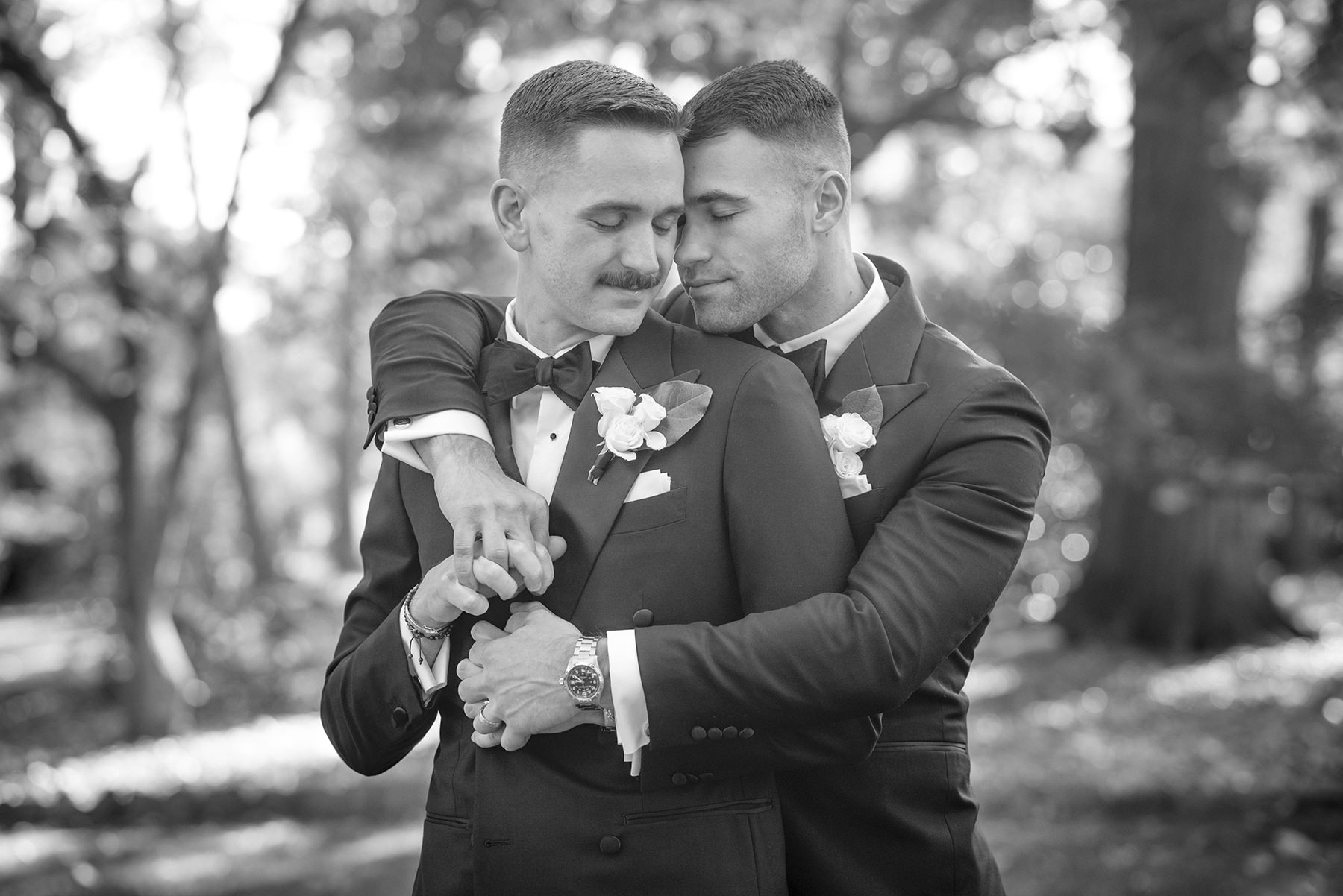 Two grooms embrace during an elegant wedding portrait at VanLandringham Estate in Charlotte, NC, surrounded by the estate's wooded grounds.