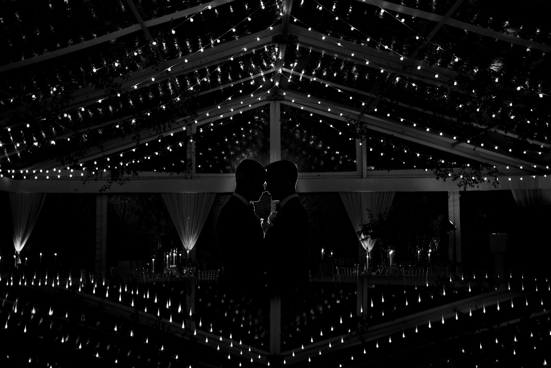 Two grooms share a romantic moment beneath a canopy of glowing string lights at their elegant VanLandringham Estate wedding reception in Charlotte, NC.